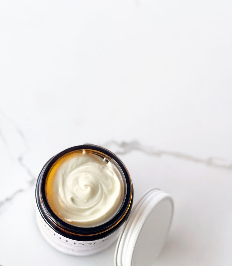 May include: A close-up of a white cream in a glass jar with a brown lid. The cream is swirled and has a smooth texture.