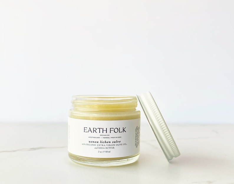 May include: A small glass jar of Earth Folk usnea lichen salve with a white label and a silver lid. The label includes the text "usnea lichen salve with ORGANIC EXTRA VIRGIN OLIVE OIL and SHEA BUTTER". The jar contains 2 oz / 60 ml of salve.