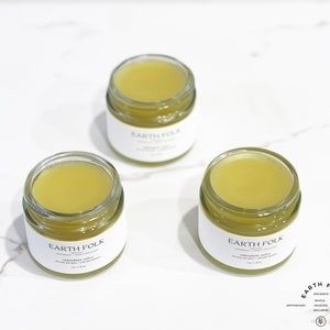Peut inclure: Trois petits pots en verre avec des &eacute;tiquettes blanches sur lesquelles est inscrit "EARTH FOLK" et contenant un baume jaune. Les pots sont pos&eacute;s sur une surface en marbre blanc.