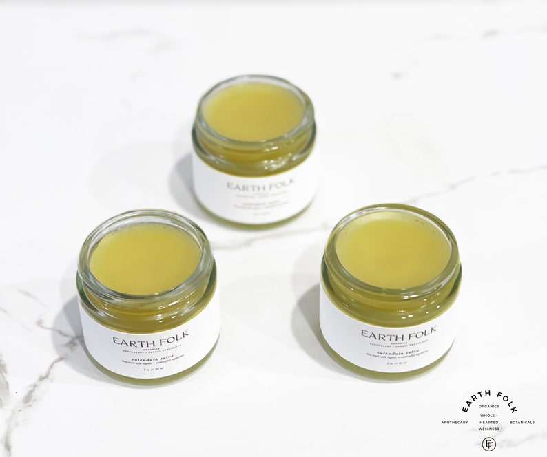 Peut inclure: Trois petits pots en verre avec des &eacute;tiquettes blanches sur lesquelles est inscrit "EARTH FOLK" et contenant un baume jaune. Les pots sont pos&eacute;s sur une surface en marbre blanc.