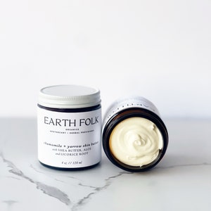 May include: Two glass jars of chamomile and yarrow skin butter with a white lid. The jar on the left has a label that reads "EARTH FOLK ORGANICS APOTHECARY HERBAL PROVISIONS chamomile & yarrow skin butter with SHEA BUTTER, ALOE and LICORICE ROOT 4 oz // 120 ml".