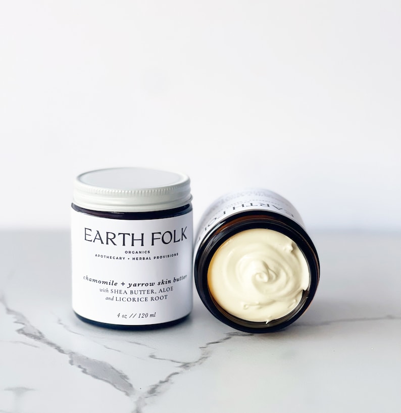 May include: Two glass jars of chamomile and yarrow skin butter with a white lid. The jar on the left has a label that reads "EARTH FOLK ORGANICS APOTHECARY HERBAL PROVISIONS chamomile & yarrow skin butter with SHEA BUTTER, ALOE and LICORICE ROOT 4 oz // 120 ml".