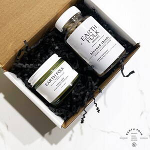 May include: A white box with black shredded paper inside. The box contains a small jar of green arnica salve and a bottle of brown capsules labeled "Earth Folk Organics Blessed Thistle Organic All Natural Non GMO Dietary Supplement 38 500mg Vegan Capsules 1-2 capsules daily".