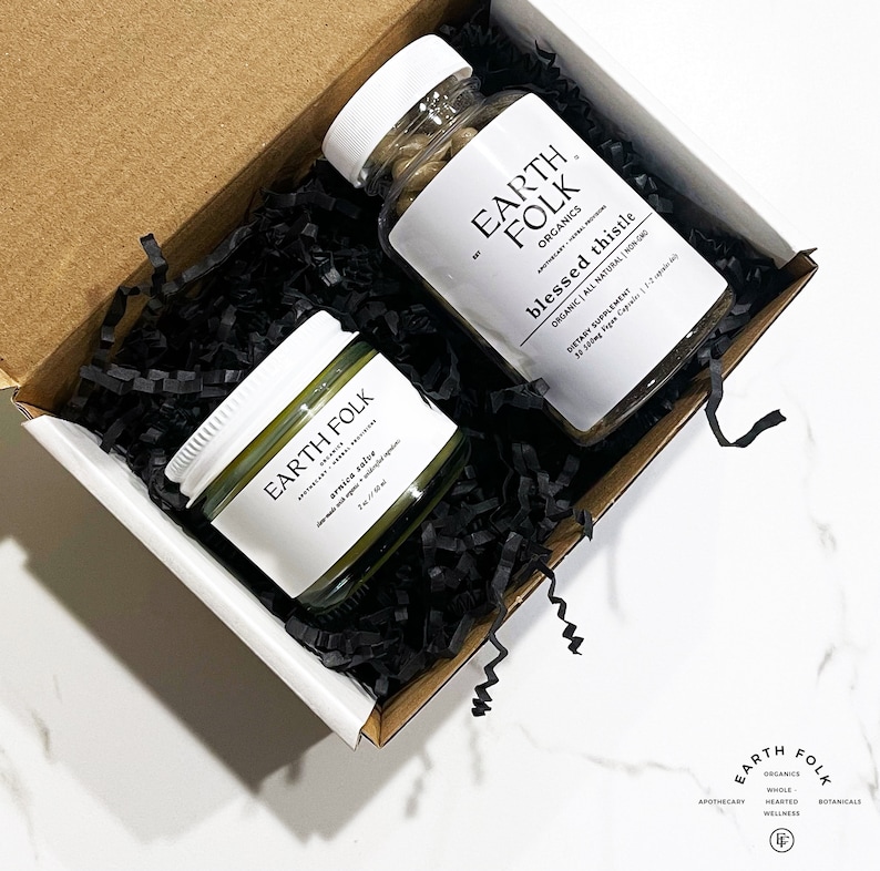 May include: A white box with black shredded paper inside. The box contains a small jar of green arnica salve and a bottle of brown capsules labeled "Earth Folk Organics Blessed Thistle Organic All Natural Non GMO Dietary Supplement 38 500mg Vegan Capsules 1-2 capsules daily".