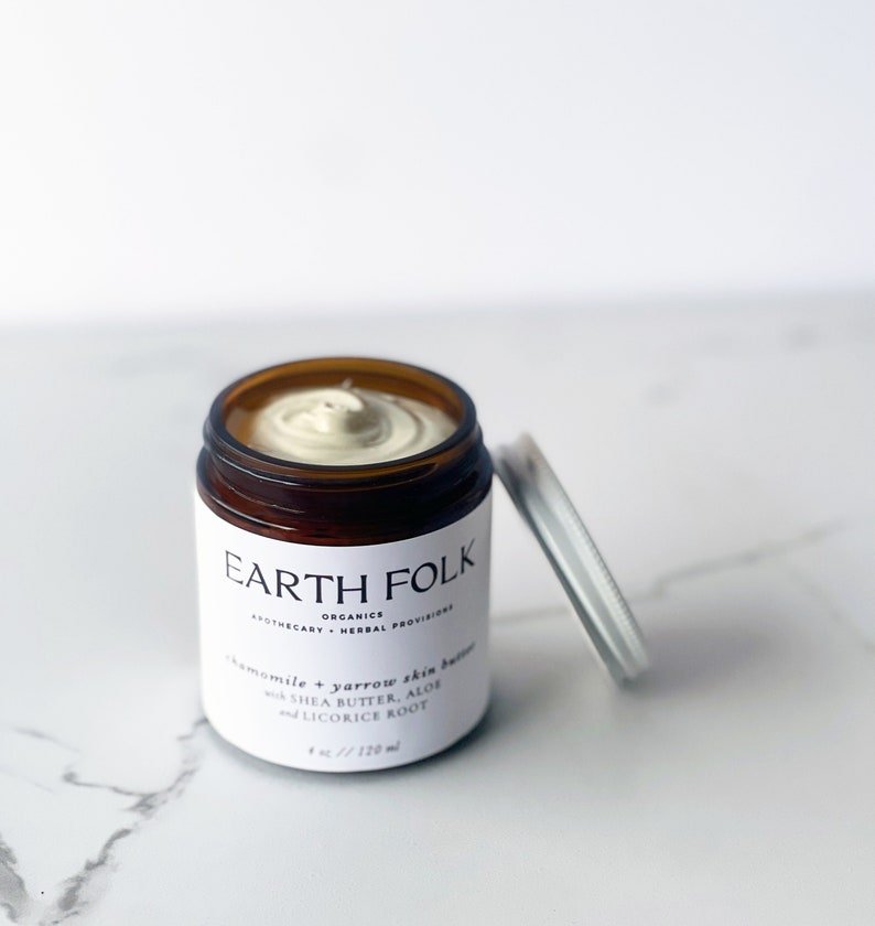 May include: A brown glass jar with a white label that reads "EARTH FOLK" and "chamomile & yarrow skin butter with shea butter, aloe and licorice root". The jar is open and contains a light beige cream.