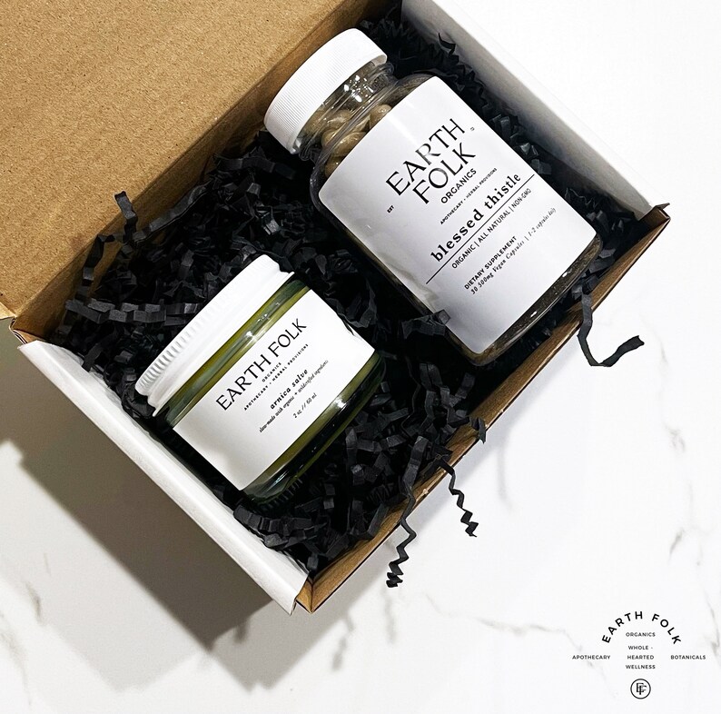 May include: A white box with black shredded paper inside. The box contains a small jar of green arnica salve and a bottle of brown capsules labeled "Earth Folk Organics Blessed Thistle Organic All Natural Non GMO Dietary Supplement 38 500mg Vegan Capsules 1-2 capsules daily".