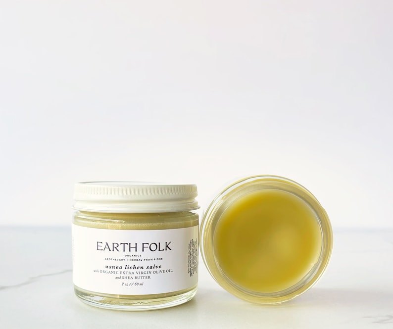 May include: Two glass jars of Earth Folk usnea lichen salve. One jar is closed with a white lid, the other is open, revealing a yellow balm. The label reads "usnea lichen salve with organic extra virgin olive oil and shea butter."