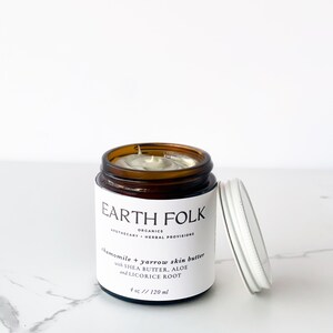 May include: A brown glass jar with a white lid containing chamomile and yarrow skin butter. The jar is labeled "EARTH FOLK ORGANICS APOTHECARY - HERBAL PROVISIONS chamomile & yarrow skin butter with SHEA BUTTER, ALOE and LICORICE ROOT 4 oz // 120 ml".