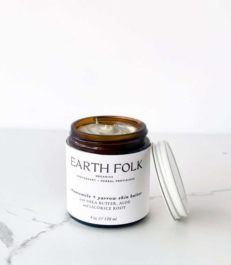 May include: A brown glass jar with a white lid containing chamomile and yarrow skin butter. The jar is labeled "EARTH FOLK ORGANICS APOTHECARY - HERBAL PROVISIONS chamomile & yarrow skin butter with SHEA BUTTER, ALOE and LICORICE ROOT 4 oz // 120 ml".