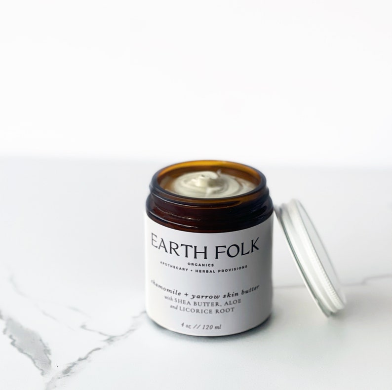 May include: A brown glass jar with a white label that reads "EARTH FOLK ORGANICS APOTHECARY + HERBAL PROVISIONS chamomile + yarrow skin butter with SHEA BUTTER, ALOE and LICORICE ROOT 4 oz / 120 ml". The jar is partially open and contains a white cream.