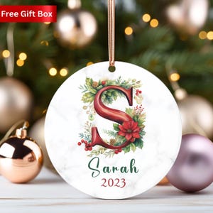 May include: Round Christmas ornament with a marble-like background. The ornament features a large, red letter "S" surrounded by greenery and a red poinsettia. The name "Sarah" and the year "2023" are printed below.