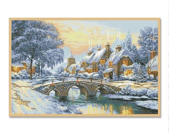 May include: A framed cross-stitch depicting a winter village scene. A stone bridge arches over a river, leading to snow-covered cottages with lit windows. The sky transitions from yellow to blue, with snow-laden trees.