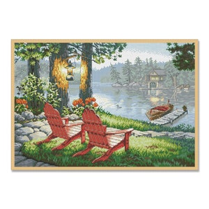 May include: Two red Adirondack chairs sit on a grassy lakeside patio with a wooden jetty and a small boat in the water. The scene is framed by trees and a cabin in the distance.