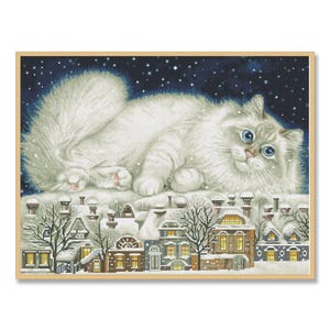 Op de afbeelding: Kruissteekkunstwerk van een pluizige witte kat met blauwe ogen, liggend op een besneeuwd dak. Daaronder een rij met sneeuwbedekte huizen met verlichte ramen en schoorstenen. De achtergrond is een donkerblauwe nachthemel met sneeuw en sterren. Gekaderd in licht hout.