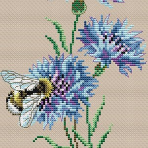 May include: Cross-stitch design of blue and purple flowers with green stems and leaves. A bee is positioned on one of the flowers. The background is a light tan color. The design is detailed and appears to be handmade.