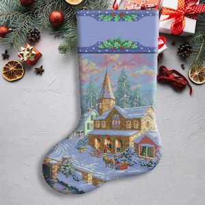 Könnte beinhalten: Ein blauer und weißer Weihnachtssocken mit einer verschneiten Dorfszene. Der Socken zeigt eine Kirche mit einem Kirchturm, Häuser und Bäume. Die Szene spielt vor einem rosa und violetten Himmel.