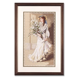 Könnte beinhalten: Eine gerahmte Kreuzstich-Stickerei, die eine Frau in einem weißen Kleid und einem Strohhut zeigt, die einen Strauß weißer Blumen hält.