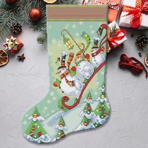 Puede incluir: Una media de Navidad verde con una escena de invierno de tres muñecos de nieve deslizándose por una colina. Los muñecos de nieve llevan sombreros y bufandas y sonríen. Hay pinos verdes a ambos lados de la colina. La media tiene un puño rojo y blanco.