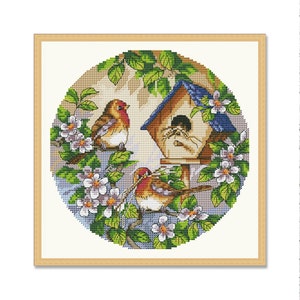 May include: A cross-stitch pattern featuring a birdhouse with a blue roof and two birds perched on branches. The birdhouse is surrounded by green leaves and pink flowers.