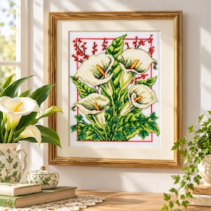 May include: A framed cross-stitch embroidery featuring white calla lilies with yellow spadices, green foliage, and small red berries, enclosed within a thin red rectangular border, set inside a gold-toned frame with a cream-colored mat.