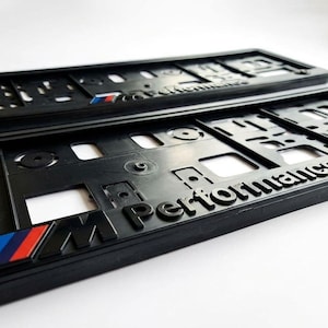 Può includere: Due portatarga neri con la scritta "Performance" in rilievo. Le cornici hanno un design a griglia e il logo "M" in blu, rosso e bianco. Per un veicolo.