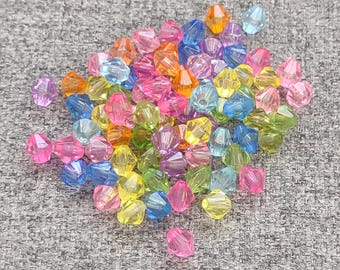 May include: A pile of colorful faceted beads in shades of pink, orange, yellow, green, blue, and purple. The beads are scattered on a gray textured surface. These beads are suitable for jewelry making and crafting.
