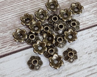 May include: A collection of antique bronze-colored flower-shaped bead caps. These small, decorative metal components feature a detailed floral design with a central hole for stringing. The bead caps are scattered on a light wood-grain surface.