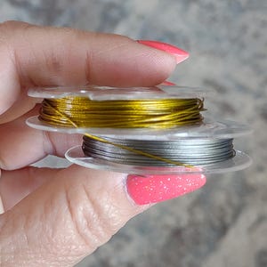 May include: Two spools of metallic wire in gold and silver. The wire is tightly wound on clear plastic spools. The gold wire is on top, and the silver wire is below. The spools are held by a person with pink nail polish.