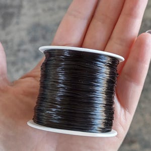 May include: A spool of black elastic cord, wound around a white plastic bobbin. The cord is thin and flexible, and it is commonly used for jewelry making and crafts.
