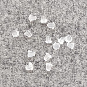 May include: A close-up of a group of clear plastic earring backs scattered on a gray fabric surface.