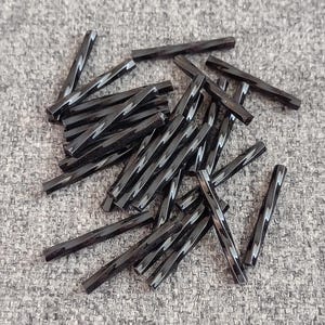 May include: A collection of small, black, twisted glass rods. Each rod has a glossy finish and a rectangular shape with a spiral design. The rods are scattered on a textured gray surface.