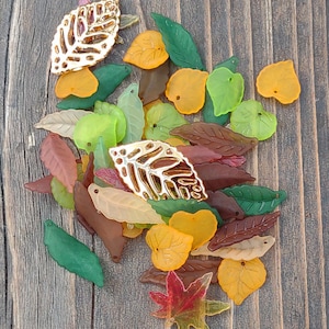 May include: A collection of colorful leaf-shaped embellishments in various shades of green, yellow, brown, and red. Some leaves have a metallic gold finish. The leaves are scattered on a wooden surface, creating a fall-themed aesthetic.