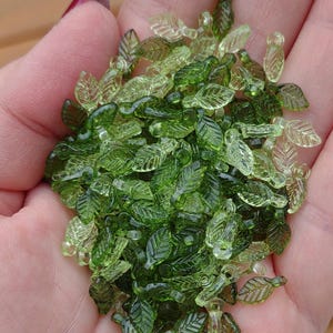 May include: A collection of small, translucent green glass leaf beads. The beads vary in shades of green, from light to dark, and are intricately detailed with leaf veins. These decorative beads are ideal for jewelry making and craft projects.