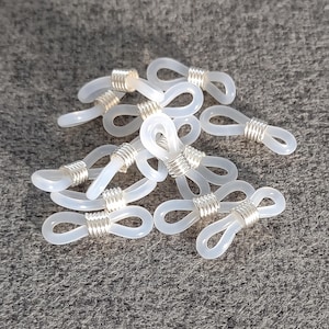 May include: A collection of clear, flexible eyeglass retainers. Each retainer features a loop design with a silver-colored metal coil wrapped around the center. The retainers are scattered on a gray surface.