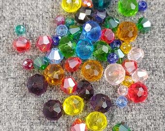 May include: A collection of faceted beads in various colors, including red, yellow, green, blue, and purple. The beads are scattered on a gray fabric surface. The beads vary in size and shape, suitable for jewelry making.