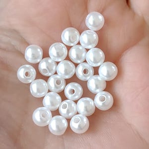 May include: A collection of small, round, white beads with a glossy finish. Each bead has a central hole, suggesting they are designed for stringing. The beads are held in a hand, showcasing their size and color.