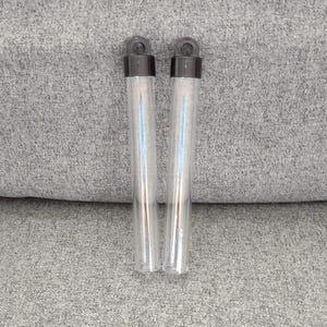 May include: Two clear glass vials with black caps. The vials are cylindrical and appear to be empty. Each vial has a small loop at the top. The vials are resting on a gray, textured surface.