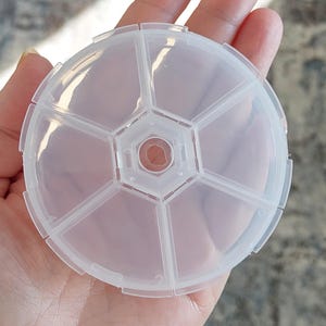 May include: A clear plastic round pill organizer with seven compartments. The organizer is held in a hand.