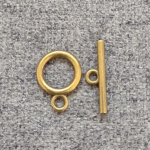 May include: A gold-colored toggle clasp, featuring a circular ring with a small loop and a straight bar with a loop. The clasp is designed for jewelry making and is shown against a gray textured background.