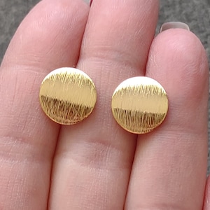 May include: Two gold-colored, round stud earrings. Each earring has a textured, brushed metal finish. The earrings are displayed against a blurred background, held by a person's fingers. The earrings are simple and elegant.