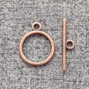 May include: A copper-colored toggle clasp set, featuring a circular ring and a straight bar with a small loop. The clasp is designed for jewelry making and is displayed against a textured gray background.