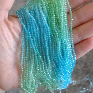 May include: Strands of small, faceted beads in shades of blue and green. The beads are held in a hand, with the colors blending together. The beads appear to be strung together, possibly for jewelry making or other crafts.