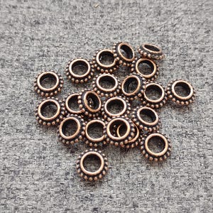 May include: A collection of copper-colored metal beads. Each bead is circular with a central hole and a textured, dotted outer edge. The beads are scattered on a gray, textured surface.