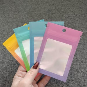May include: A collection of colorful, resealable plastic pouches in yellow, blue, and pink. Each pouch has a clear window and a small hole at the top. The pouches are held in a hand against a gray background.