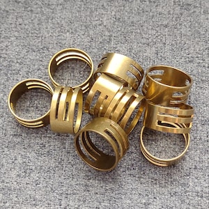 May include: A collection of gold-colored metal rings with various designs. Some rings have a solid band, while others feature vertical cutouts. The rings are scattered on a gray textured surface, showcasing their metallic sheen.