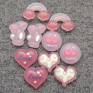 May include: A collection of pink resin embellishments, including rainbows, bunnies, cherries, and hearts. The hearts feature the word "Love" and a patterned design. These decorative pieces are ideal for crafting and scrapbooking projects.