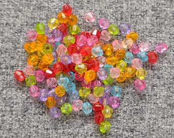 May include: A pile of colorful, faceted beads in various shades of red, orange, yellow, green, blue, pink, and purple. The beads are scattered on a gray, textured surface, showcasing their translucent quality and diverse hues.