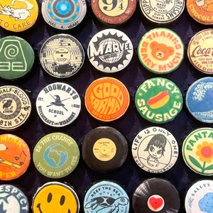 Könnte beinhalten: Eine Sammlung runder, farbenfroher Ansteckbuttons mit verschiedenen Designs und Texten. Die Buttons zeigen Bilder wie eine Katze, ein Herz, einen Bären, einen Hotdog und die Texte "Hogwarts" und "Marvel".