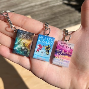 May include: Three miniature book keychains featuring covers for Percy Jackson by Rick Riordan, Heated Rivalry by Rachel Reid, and Our Perfect Storm by Carley Fortune. Each rectangular charm is attached to a silver metal keyring with a chain link.