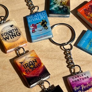 Op de afbeelding: Een verzameling miniatuur boek-sleutelhangers met diverse titels zoals Fourth Wing, Heated Rivalry, Iron Flame en Our Perfect Storm. Elke sleutelhanger bestaat uit een kleine rechthoekige boekreplica bevestigd aan een zilveren metalen ring.